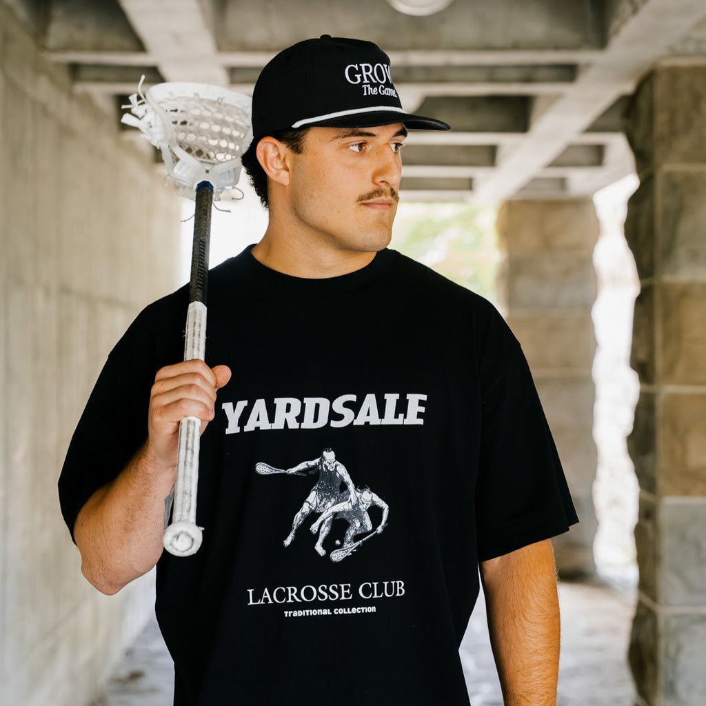 Man holding a lacrosse stick wearing a black t-shirt with 'Yardsale Lacrosse Club' design.