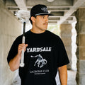 Man holding a lacrosse stick wearing a black t-shirt with 'Yardsale Lacrosse Club' design.