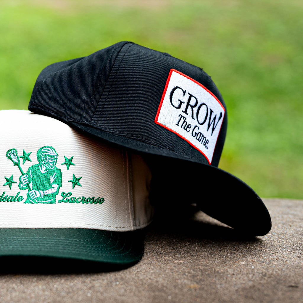 Two snapback caps displayed outdoors on a stone surface with a blurred green grass background. The front cap is white with a dark green brim and green Yardstate Lacrosse embroidery.