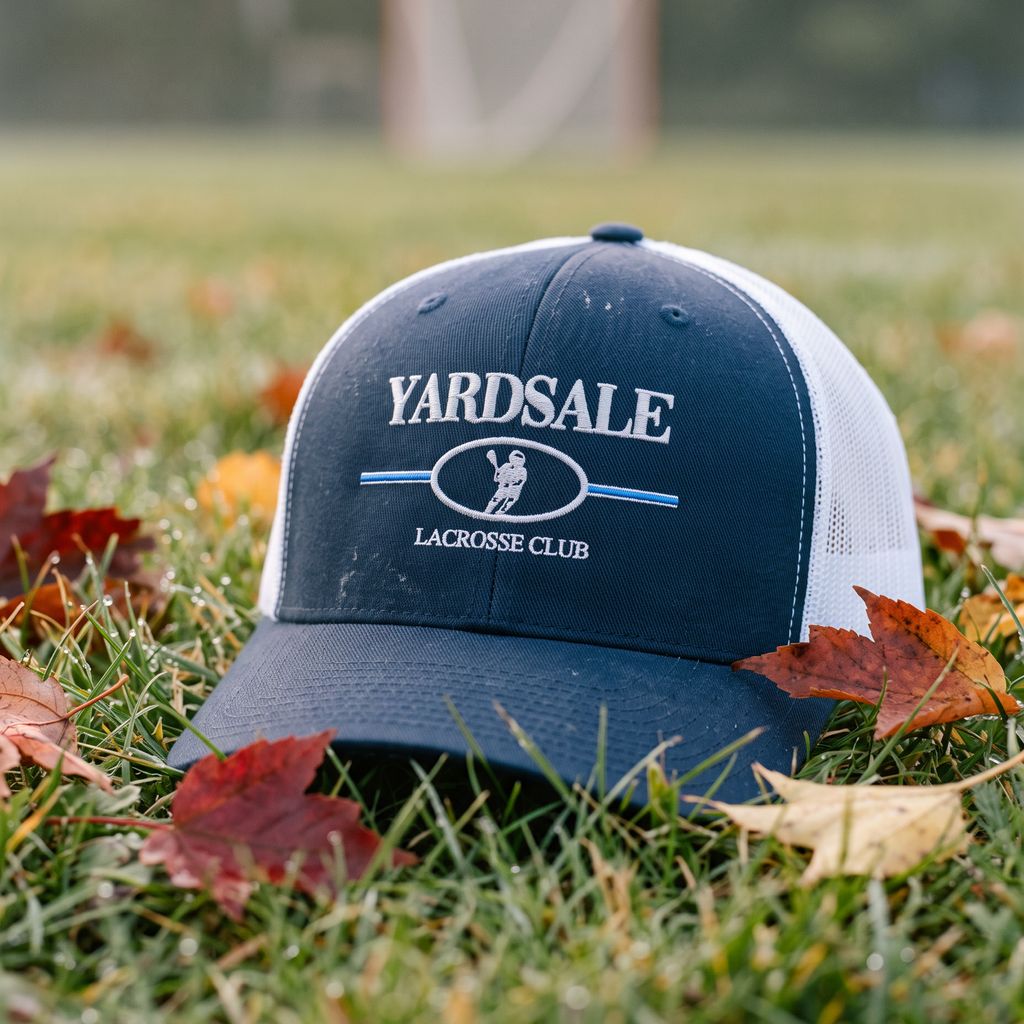 A navy blue and white trucker hat with 'YARDSALE LACROSSE CLUB' embroidered on the front, resting on green grass scattered with autumn leaves in an outdoor field.