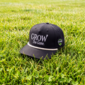 A black baseball cap with white embroidered text reading 'GROW The Game.' and a small side patch, placed on green grass outdoors.