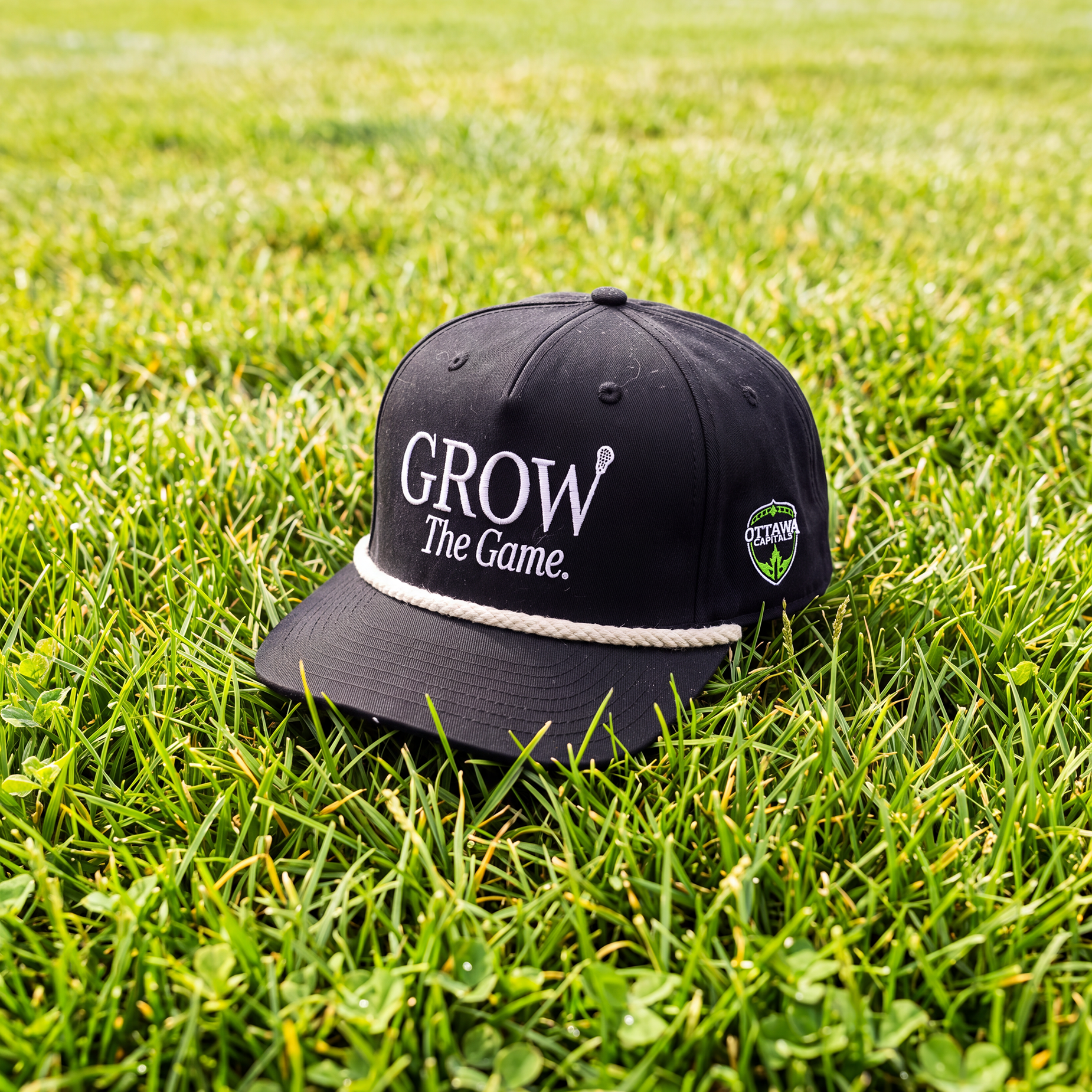 A black baseball cap with white embroidered text reading 'GROW The Game.' and a small side patch, placed on green grass outdoors.