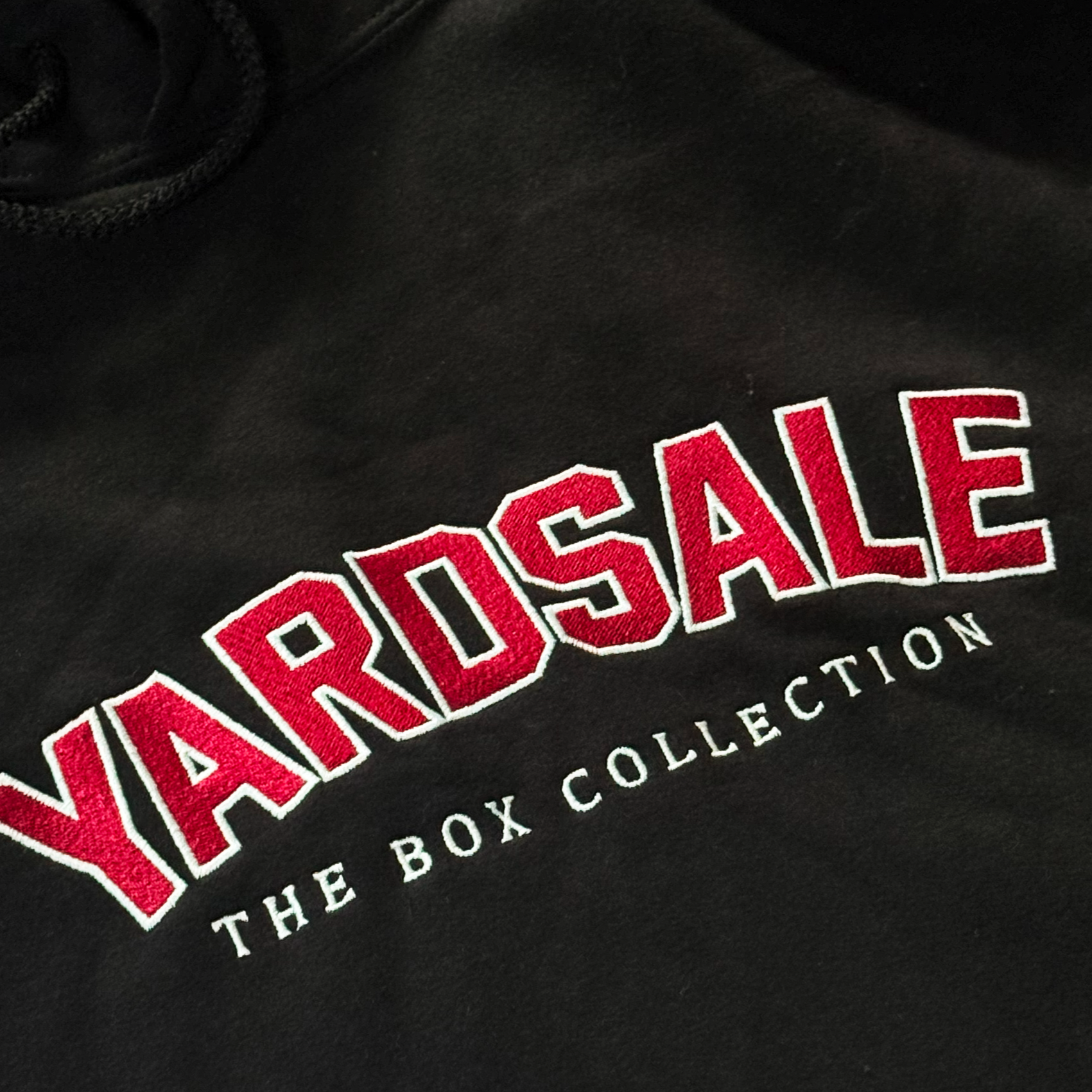 Black hoodie with 'YARDSALE THE BOX COLLECTION' text on a white surface.
