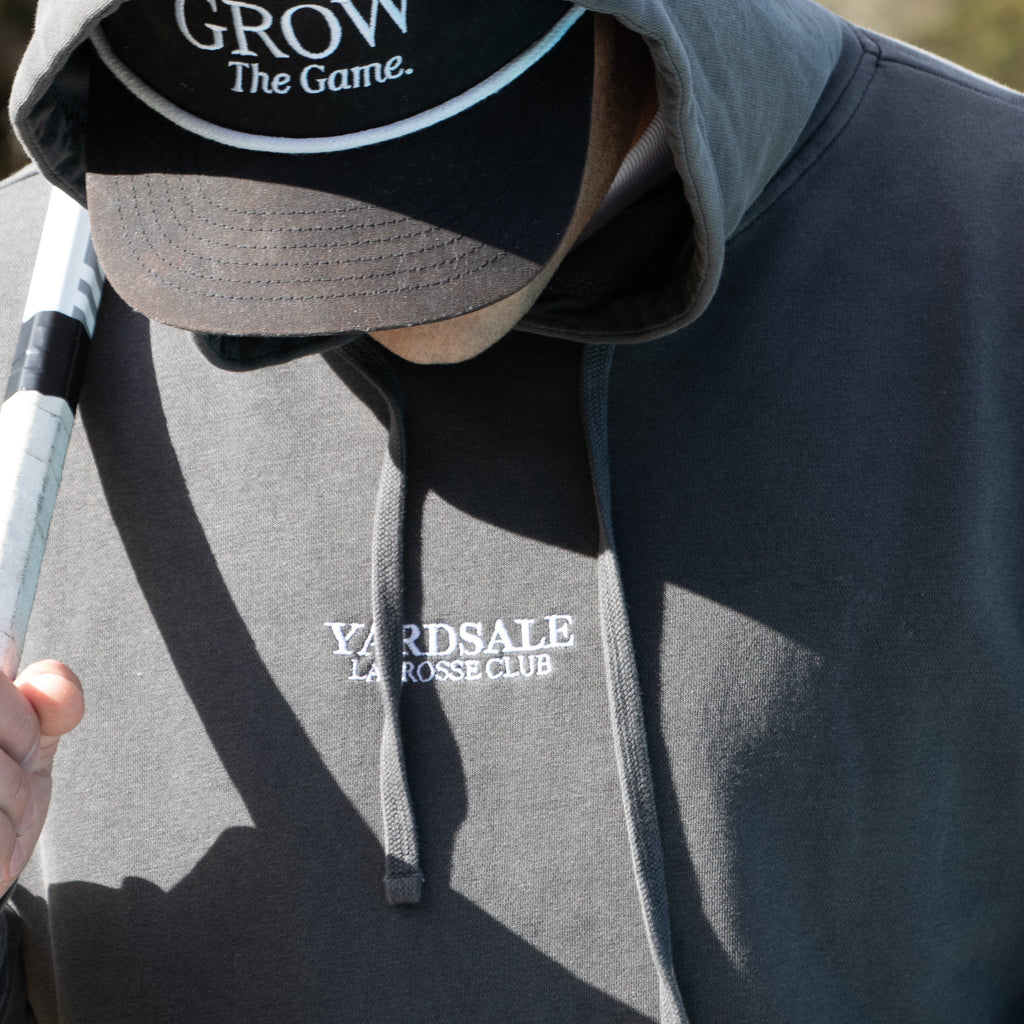Person wearing a hoodie and cap with lacrosse equipment, outdoors.