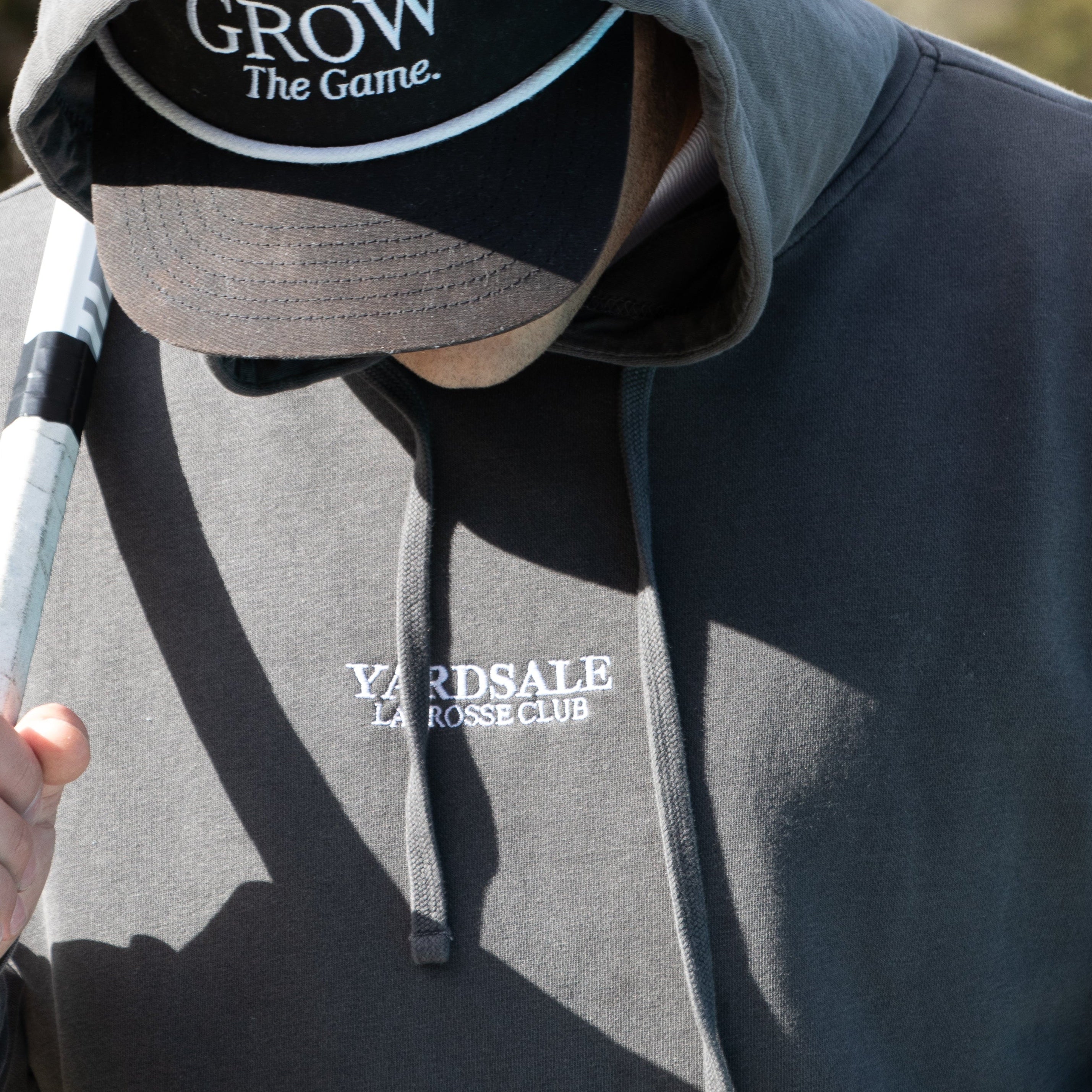 Person wearing a hoodie and cap with lacrosse equipment, outdoors.