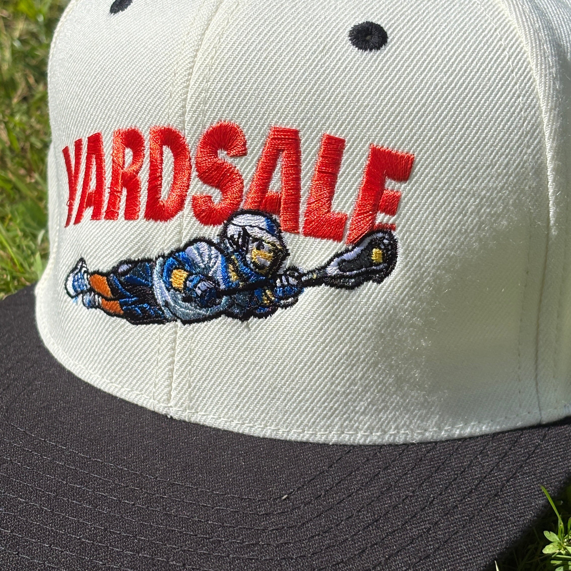 A structured snapback cap with an off-white crown and a black flat brim, resting on green grass. The front of the cap features 'YARDSALE' embroidered in red across the forehead, with a cartoon catcher in a blue and orange uniform below it.