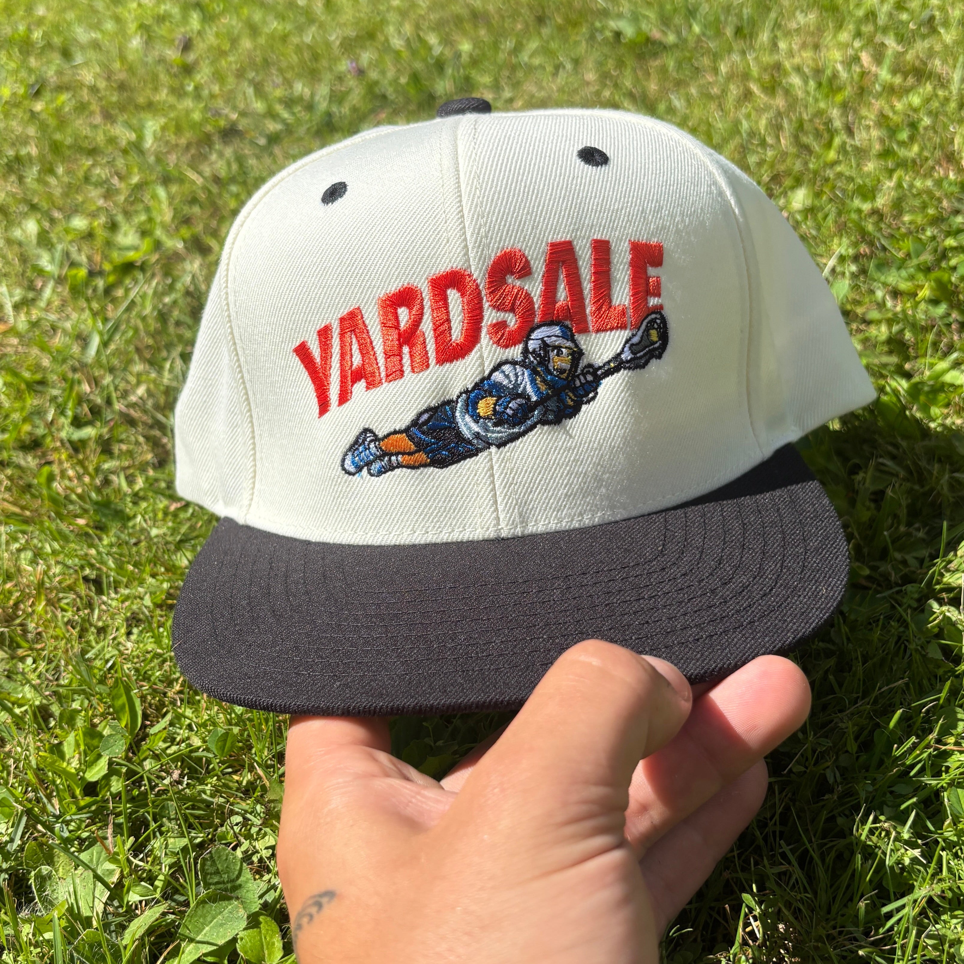 A white and black snapback cap held by a hand outdoors on a grassy lawn. The cap features 'YARDSALE' embroidered in red on the front, with a blue and orange cartoon character below it.