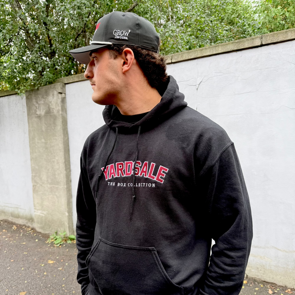 Man wearing a black hoodie with 'Yard Sale' text, standing outdoors against a wall.