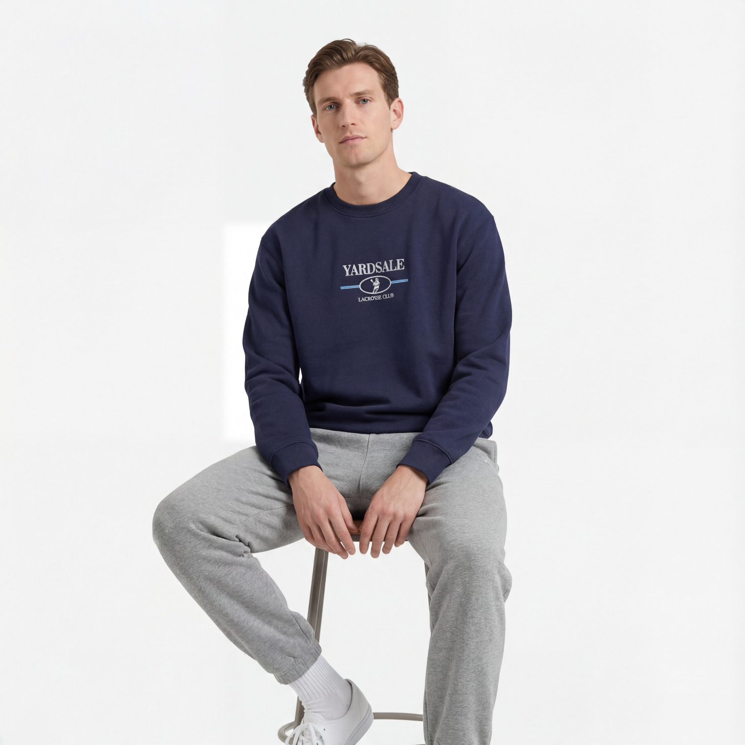 Man sitting on a stool wearing a navy sweatshirt with a logo and gray pants on a white background