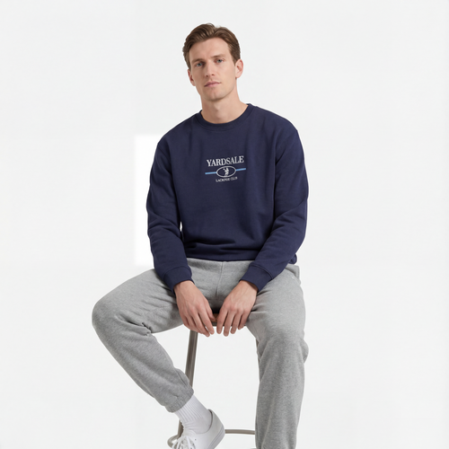Man sitting on a stool wearing a navy sweatshirt with a logo and gray pants on a white background