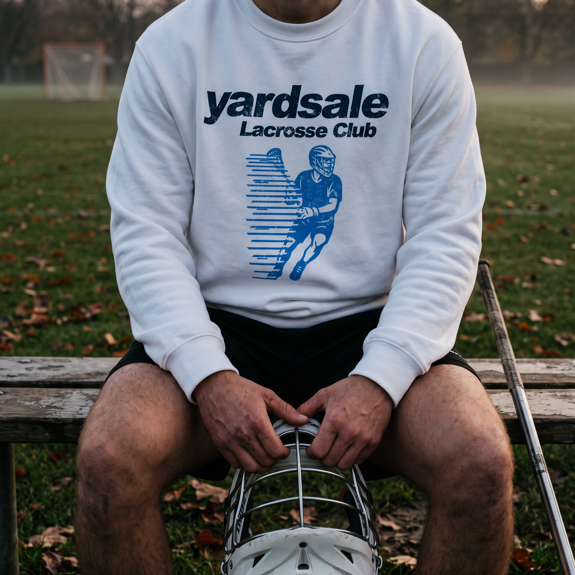 A man sitting on a wooden bench outdoors on a grassy field, wearing a white crewneck sweatshirt with a blue 'yardsale Lacrosse Club' graphic and holding a lacrosse helmet in front of his legs.
