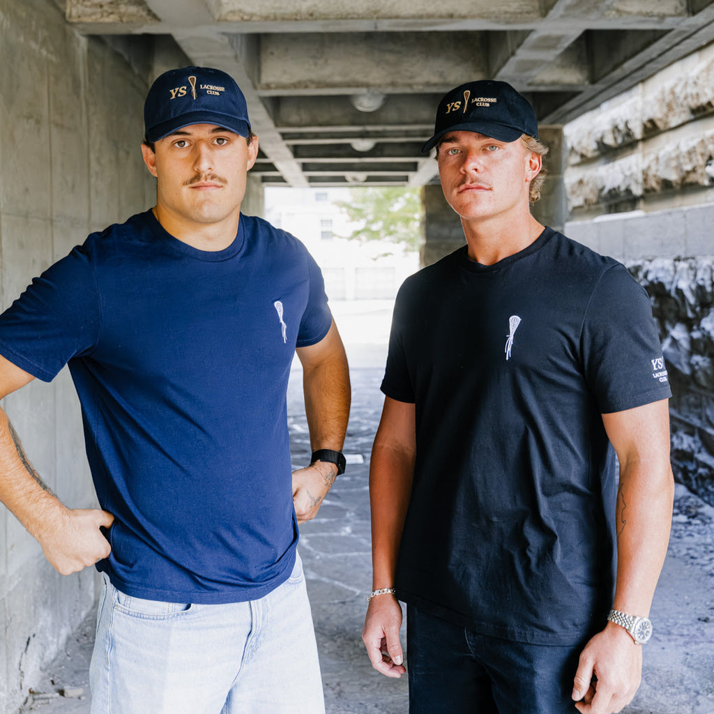 Two men standing under a concrete overpass wearing matching blue t-shirts and caps.