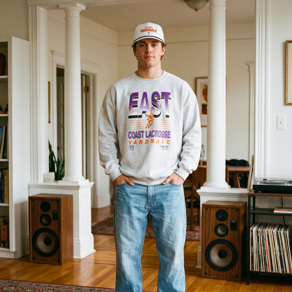 Person wearing a sweatshirt and jeans standing in a room with speakers and a rug.
