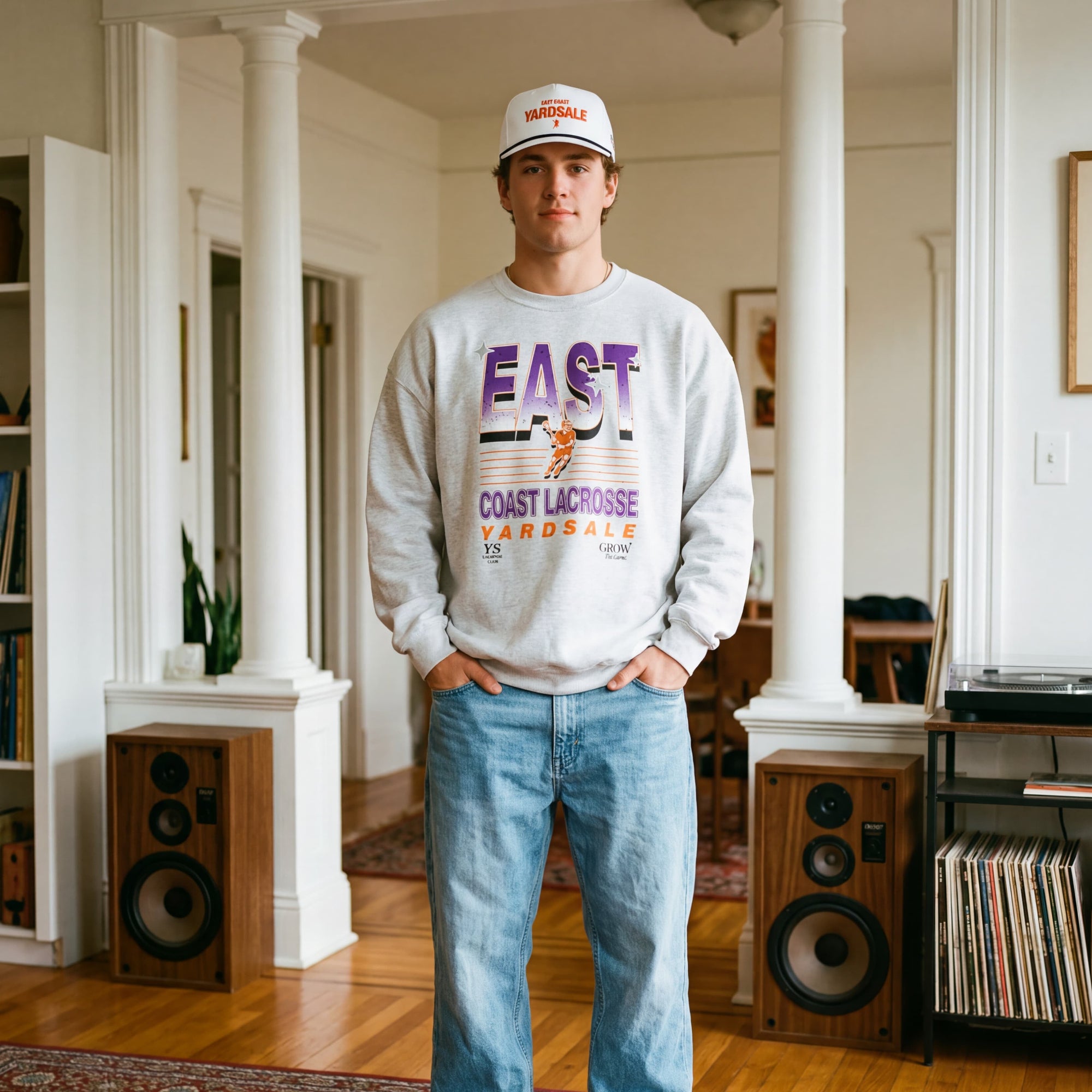 Person wearing a sweatshirt and jeans standing in a room with speakers and a rug.