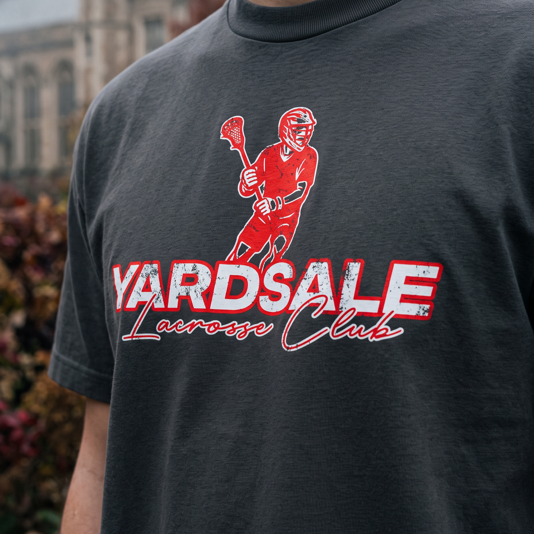 A close-up of a person wearing a dark charcoal gray t-shirt with a red and white graphic of a lacrosse player and the text 'YARDSALE Lacrosse Club'. The background shows a blurred outdoor scene with a stone building.