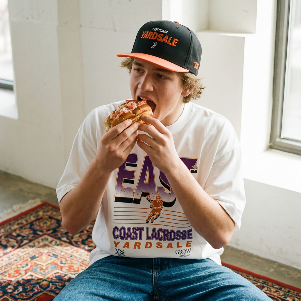 Person eating a sandwich wearing a cap and t-shirt with text, sitting on a rug.