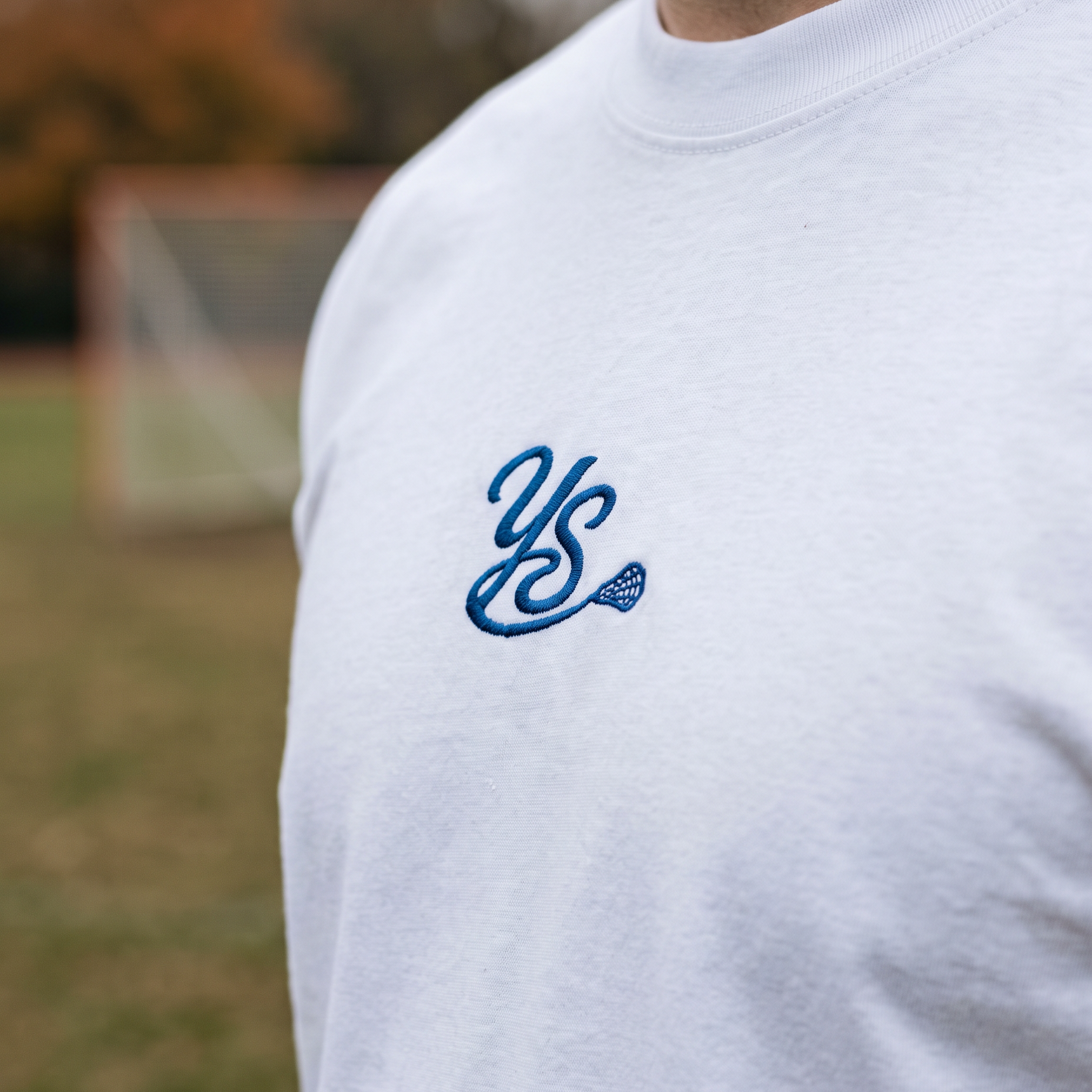 A close-up of a person wearing a white crewneck t-shirt with a blue embroidered 'YS' monogram on the chest, set against a blurred outdoor background of a sports field.