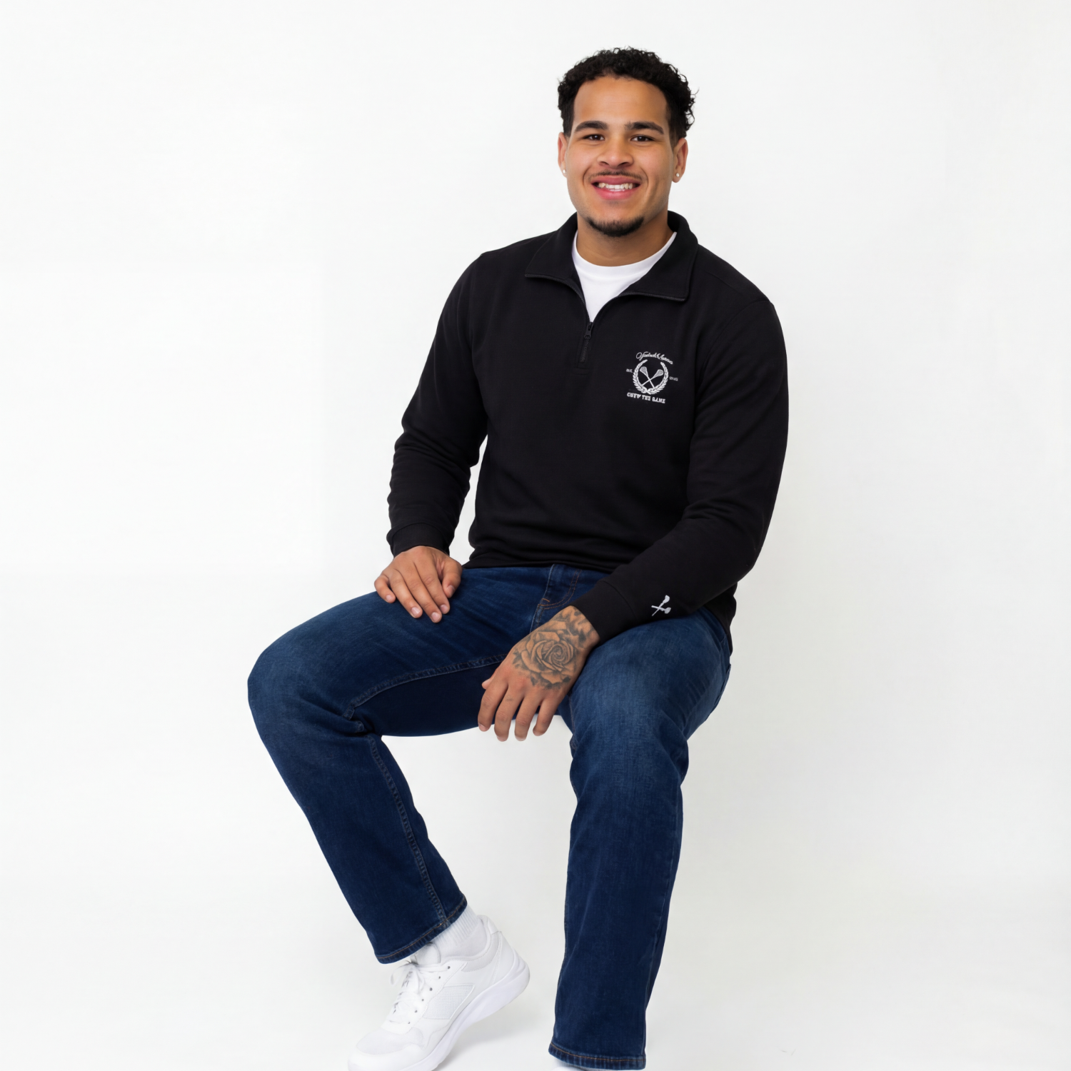 Man wearing a black sweater with a logo, blue jeans, and white sneakers on a white background