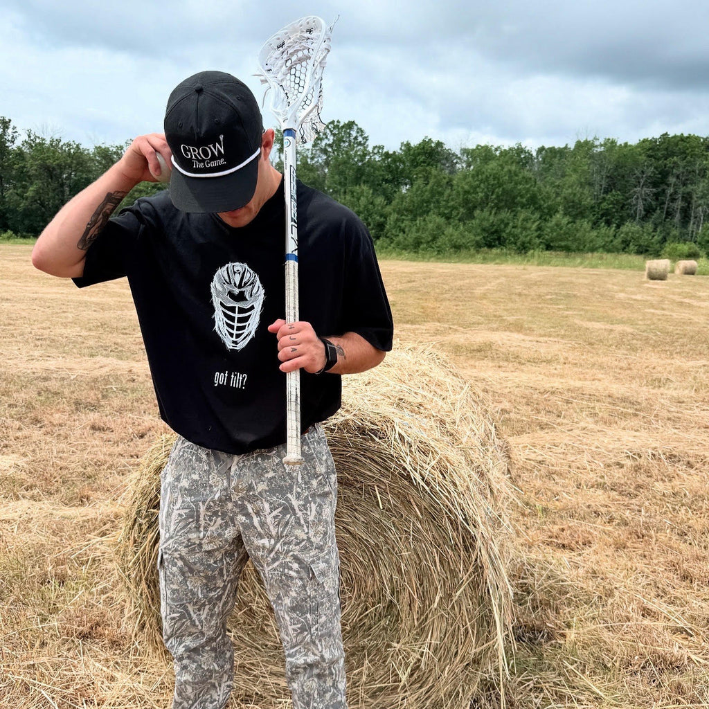 Person holding a lacrosse stick standing on a hay bale in an open field with trees in the background