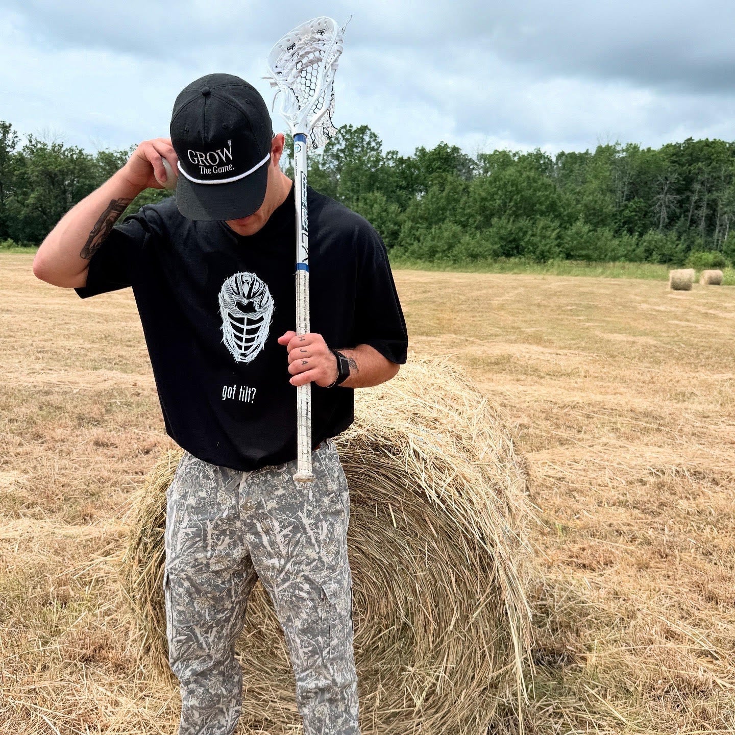 Person holding a lacrosse stick standing on a hay bale in an open field with trees in the background