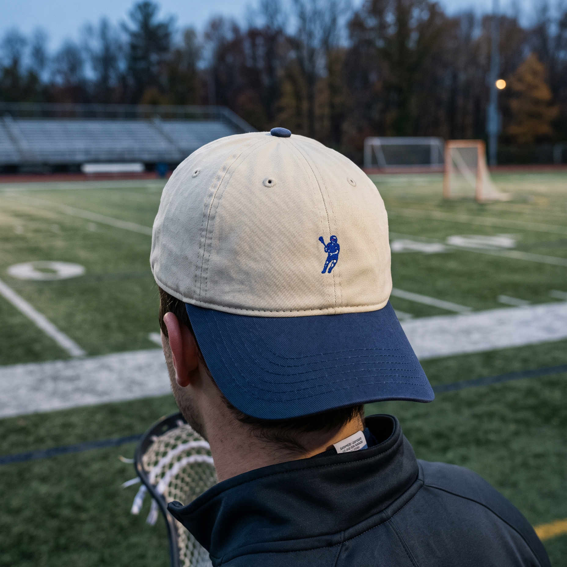 A person viewed from behind wearing a baseball cap with a cream crown and navy blue visor, featuring a small blue lacrosse player logo on the side. The background shows a blurred outdoor lacrosse field at dusk.
